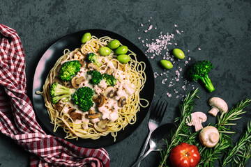 Close-up of traditional delicious italian pasta with chicken, mushrooms, broccoli, cream sauce and green olives on black plate on dark background. Top view. Dark low key photo. Flat lay composition.