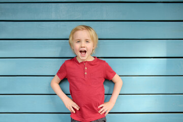 Cheerful and energetic little boy wearing red t-shirt standing on blue background. Child is joyful...