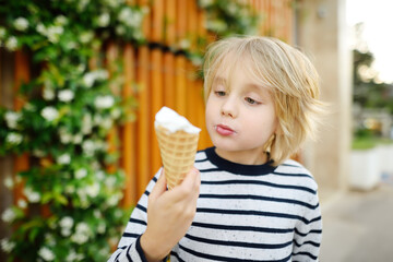 Fototapeta premium Happy little boy eating tasty ice cream cone outdoors during family stroll. Child have a snack on the go. Gelato is loved delicacy of kids. Sweets are unhealthy food for baby. Diabetes risk