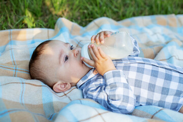 Adorable baby drinking milk from bottle while lying on blanket at picnic. Baby nutrition.