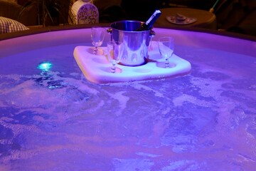 Champagne bottle in ice bucket and 4 glasses on a float in bubbling jacuzzi bath with purple lights under water - obrazy, fototapety, plakaty