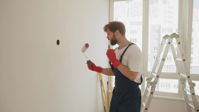 Young worker painting wall in room. Happy caucasian male singing song using roller brush as a microphone. Young man having fun during home repair and renovation. Concept of successful repair.