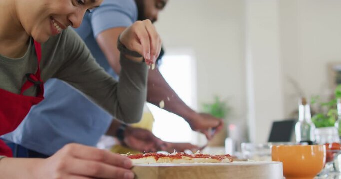 Smiling diverse female and male friends making pizza and cooking in kitchen, in slow motion