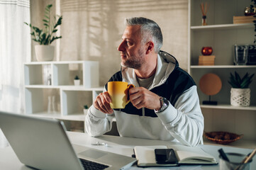 Middle aged man a laptop while working in a home office and drinking coffee