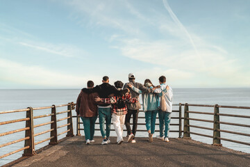 A diverse group of young friends come together in a tight embrace, facing the sea and the horizon....