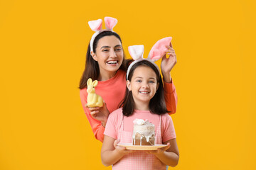 Beautiful woman and her little daughter in bunny ears with Easter cake and rabbit on yellow background