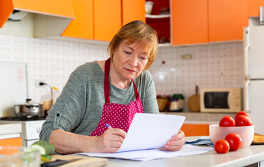 Obraz na płótnie Canvas Focused middle aged woman signing paper documents while sitting in cozy home kitchen