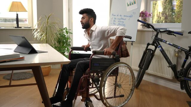 Wide shot young motivated Middle Eastern man in wheelchair making effort standing up. Portrait of handsome intelligent freelancer rehabilitating in home office indoors sighing thinking
