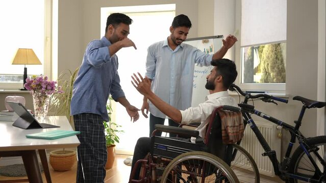 Smiling happy Middle Eastern men stacking hands with friend in wheelchair tapping shoulders talking. Positive confident young friends rejoicing startup success in home office indoors