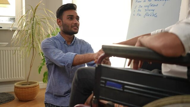 Smiling positive Middle Eastern young man sitting down on one knee taking friend hand talking. Portrait of supportive guy endorsing mate in wheelchair indoors. Rehabilitation and friendship