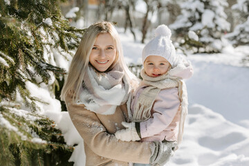 Fototapeta premium Portrait of young mom and little daughter enjoying sunny day at snowy park. Mother holding child on hands, and looking together at camera while standing in background of snowy trees