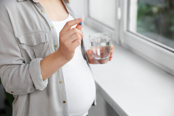 Pregnant woman holding pill and glass with water near window indoors, closeup
