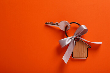 Key with trinket in shape of house and grey bow on orange background, top view. Housewarming party