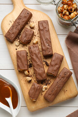 Board with delicious chocolate bars, caramel and nuts on white wooden table, flat lay