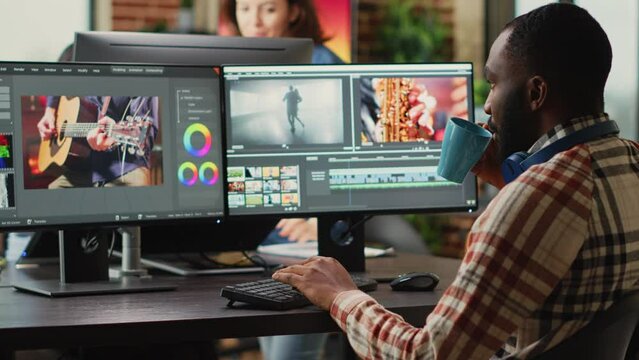 African american employee editing video content on multimedia software, using focus lighting and color grading. Male filmmaker working on movie montage edit on pc multi monitors.