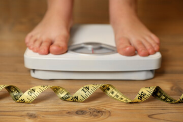 Overweight girl using scales near measuring tape on wooden floor, selective focus