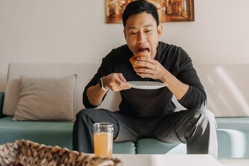 Asian man having bread and juice as breakfast on the sofa living room.