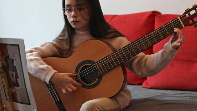 A beautiful little caucasian brunette girl with flowing long hair in glasses holds a guitar and plucks the strings with her fingers, learning notes while sitting on a sofa in the room, close-up 