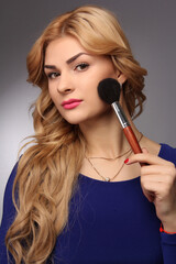 portrait of a blonde makeup artist girl with blonde hair in a blue dress with face brushes in the studio room, with makeup and hairstyle, red lips and manicure, draws