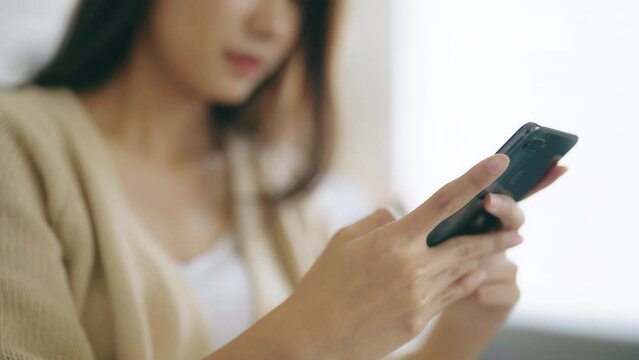 Happy young asian woman relax on comfortable couch at home, typing chat message on smartphone, smiling girl use cell phone chatting, searching information on browser wireless internet, online shopping