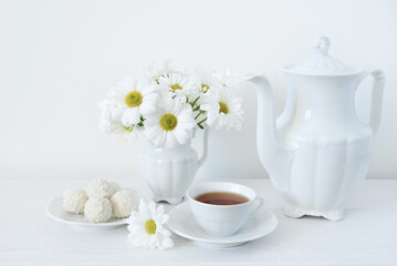Tea and flowers. A bouquet of white flowers of daisies or chamomile in a vase on the table, a cup of tea, sweets and a teapot on a white background