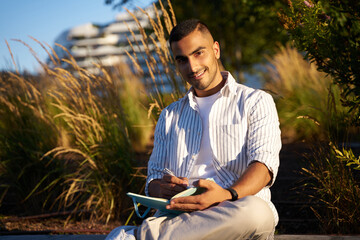 Smiling middle eastern man looking at the camera and taking notes at the notebook sitting in park 