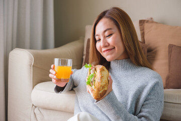Portrait image of a young woman holding and eating french baguette sandwich and orange juice at home