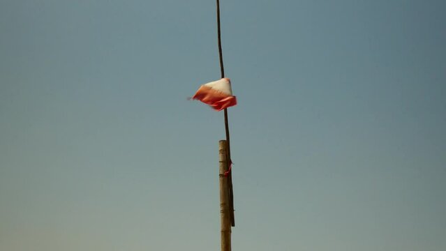 A tattered Peruvian flag waving in the wind. The flag itself is attached to a wooden pole attached to a bigger pole. The background is a blue sky.