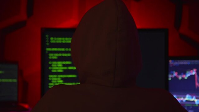 Anonymous hacker in a dark room with multiple monitors writing code.