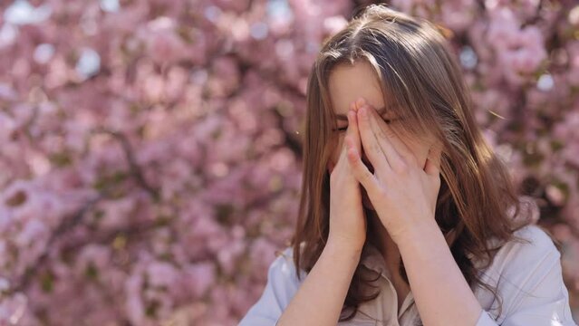 Irritated caucasian young woman standing near blooming sakura tree and scratching itchy eyes during spring time outdoors. Allergic reaction to pollen. For Advertisement of a medicinal product