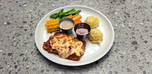 Chicken Parmigiana set of plate with mashed potato, corn slash, pepper seasoned carrot & long peas, accompanied with mushroom and barbeque sauce on a white plate. Served on top of grey terrazzo table.