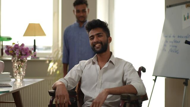 Successful young man in wheelchair with toothy smile looking away as friend standing at background in home office. Positive motivated Middle Eastern freelancer indoors with supportive mate