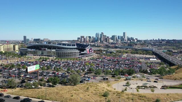 Cinematic aerial tracking view of Empower field at Mile High Stadium with lots of cars parked in parking spaces and main street highway and Downtown Denver city view, Colorado