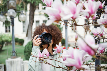 In front of the pink magnolia buds in the spring park, a girl photographer with a camera makes photos of beautiful flowers. Springtime, hobby, no allergy