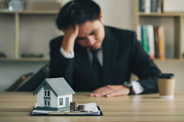 businessman sitting stressed holding his head with the house model and contract document in front....