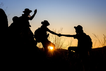 silhouette of Scout students showing help to each other Hold hands and pull each other up from the cliff to explore the beautiful forest in the evening as the sun sets. - obrazy, fototapety, plakaty