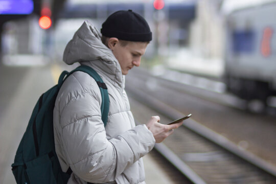 Young man using smartphone, texting, cheks the route of trip on google mapes while waiting train at railway station.