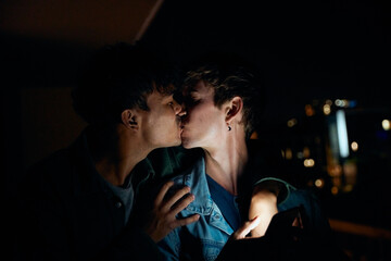 Young gay couple in casual clothing kissing while holding mobile phone on balcony at night