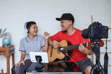 Fototapeta na wymiar Dad and son singing and playing guitar together while doing video online streaming. Asian family spending quality time at home while recording video on digital camera.