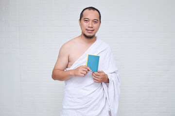 Muslim man wearing white ihram clothes and holding passport ready for Hajj or umrah, standing over...