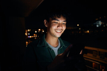 Young multiracial man in shirt smiling while using mobile phone on balcony of apartment at night