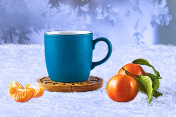 Mandarins with leaves and slices, a blue cup of coffee on a white snowy background. Winter still life with snowflakes