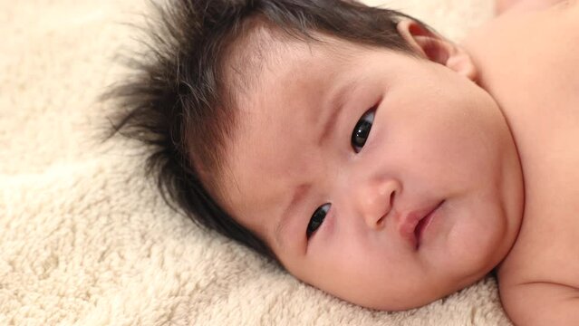 Close-up the face of asian newborn baby lying on beige bed looking camera and looks around with doubt, adorable infant early days who wakes up and open his eyes. Concept of caring for children.