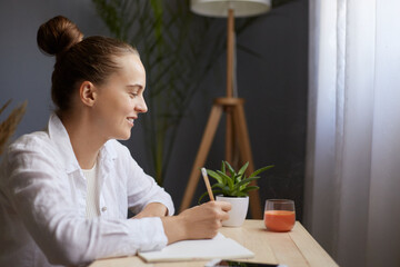 Image of smiling positive optimistic young beautiful woman writing down notes while sitting at table in office, thinking about to do list, expressing positive emotions.