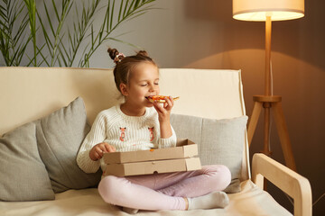 Indoor shot of cute funny child biting big slice pizza, sitting on sofa with crossed legs, enjoying...