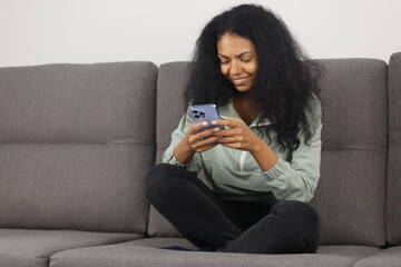 Happy black female person typing online message with modern smart phone. Cheerful POC woman sitting on couch at home and using mobile phone