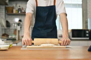 Man wearing aprons preparing homemade pastry, kneading dough with a rolling pin on wooden table in kitchen