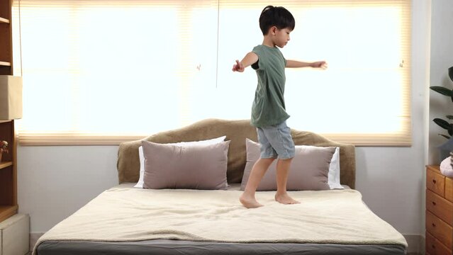 Happy asian elementary school boy jumping and dancing on bed, kid jumped and stomped around joyfully alone in bedroom without feeling tired. little boy have fun on the weekend. healthy child concept.
