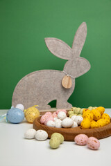 Multi-colored chocolate decorative eggs in wooden plate and gray rabbit on a green background. Happy Easter background. Front view