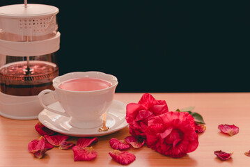 A white teacup of pink rose tea on saucer on a wooden table with two-tone red roses and falling...